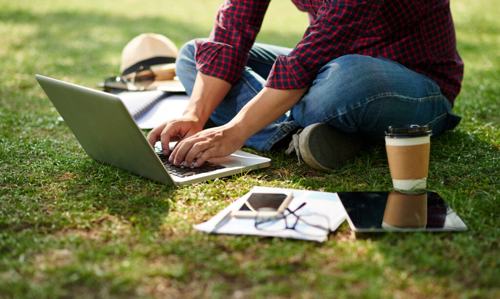 A person in a plaid shirt and jeans types on a laptop set up on a grassy field. Around the person is a coffee cup, phone, glasses, papers and other items.