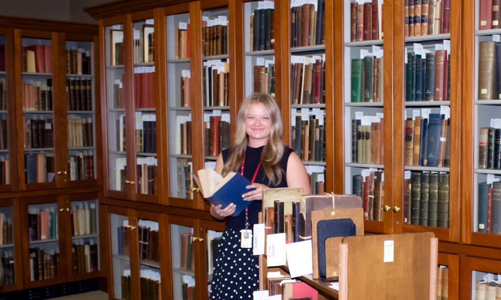 Kara Walters, a student worker at McGoogan Library, holds a book while standing before shelves and carts full of texts. 