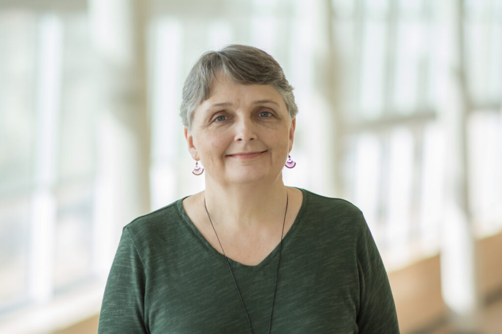 Cindy Schmidt wears a green long-sleeved top with purple fan earrings against a well-lit hallway. 